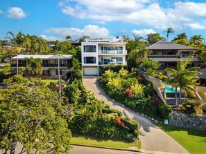 Une maison moderne à deux étages, dotée d’une allée menant à une entrée sécurisée par un portail, et entourée d’une végétation tropicale luxuriante.