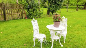 Ein weißer Sitzbereich im Freien mit Tisch und Stühlen, umgeben von üppiger Vegetation und einem Holzzaun.