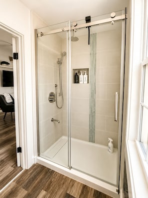 A modern bathroom with a glass-enclosed shower, a handheld showerhead, a wall-mounted faucet, and a small shelf with toiletries.