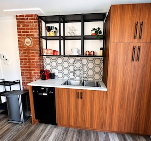 A compact kitchen with a dishwasher, a sink, a black cabinet, a wooden cabinet, and a brick wall.