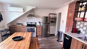 A modern kitchen with a wooden table, built-in oven, stainless steel refrigerator, and a brick accent wall. - Historic Bruce House | Hot Tub, Fire Pit &AC (Shelburne)