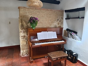 Piano, jonka päällä on nuotteja, penkki, matkalaukku ja kirjahylly täynnä kirjoja.