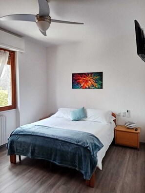 A bedroom with a bed, a nightstand, a wall-mounted TV, a ceiling fan, and a window with curtains. - Detached house, A/C, free parking, big garden, patio and Solarium with shower (Como)