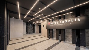 En modern hotellobby med en skylt som visar namnet ”Hotel Legalie Osaka Nipponsashi South”. Lobbyn har en stilren och modern design med en vägg av sten och ett klinkergolv.