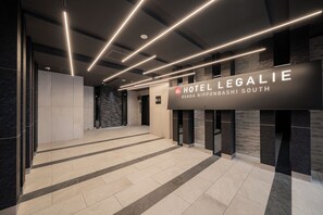 Eine moderne Hotelhalle mit einem Schild für 'Hotel Legalie Osaka Nipponsashi South', die ein klares, zeitgemäßes Design mit einer Steinwand und einem gefliesten Boden aufweist.