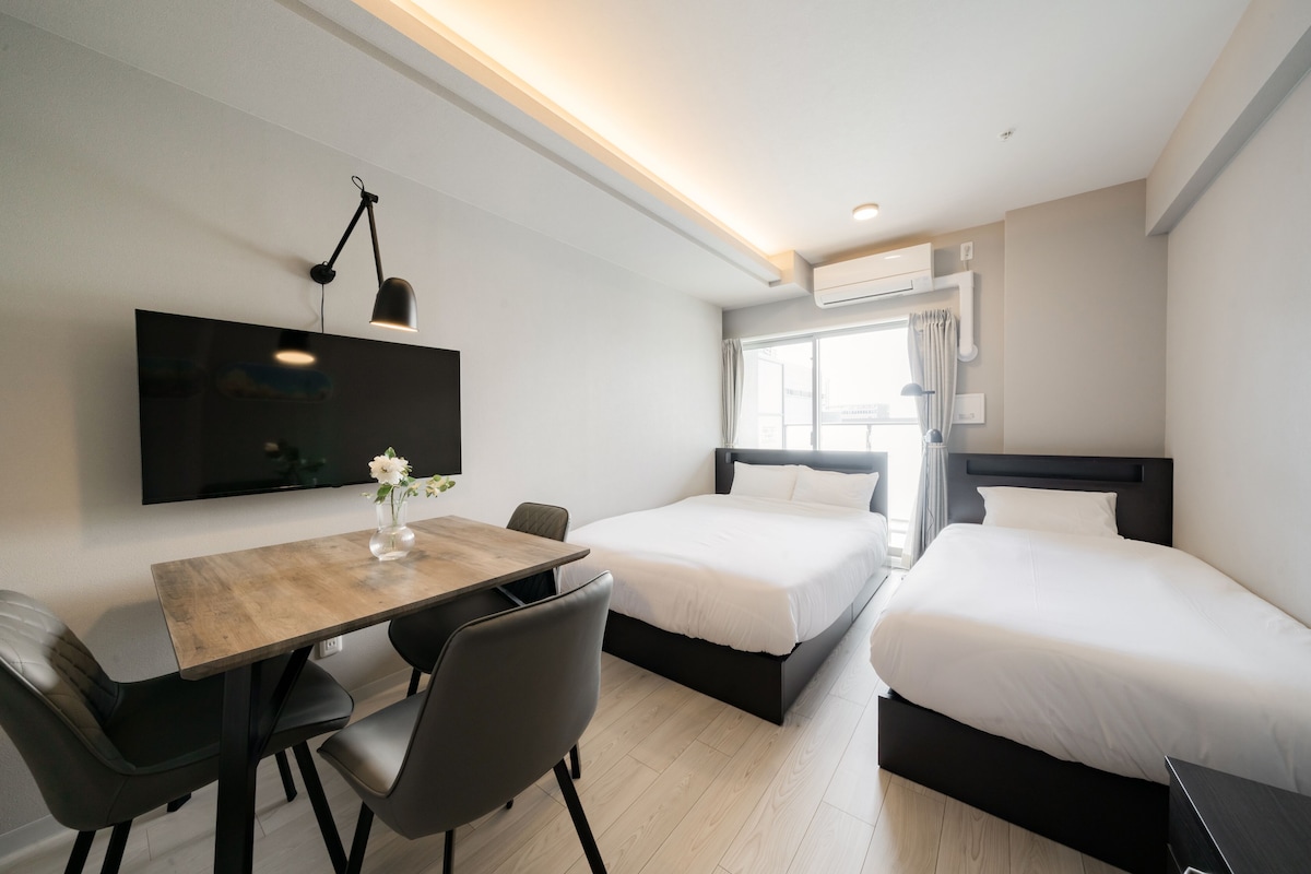 Ein modernes Hotelzimmer mit Flachbildfernseher, Esstisch aus Holz, zwei Betten und einem großen Fenster mit Vorhängen.