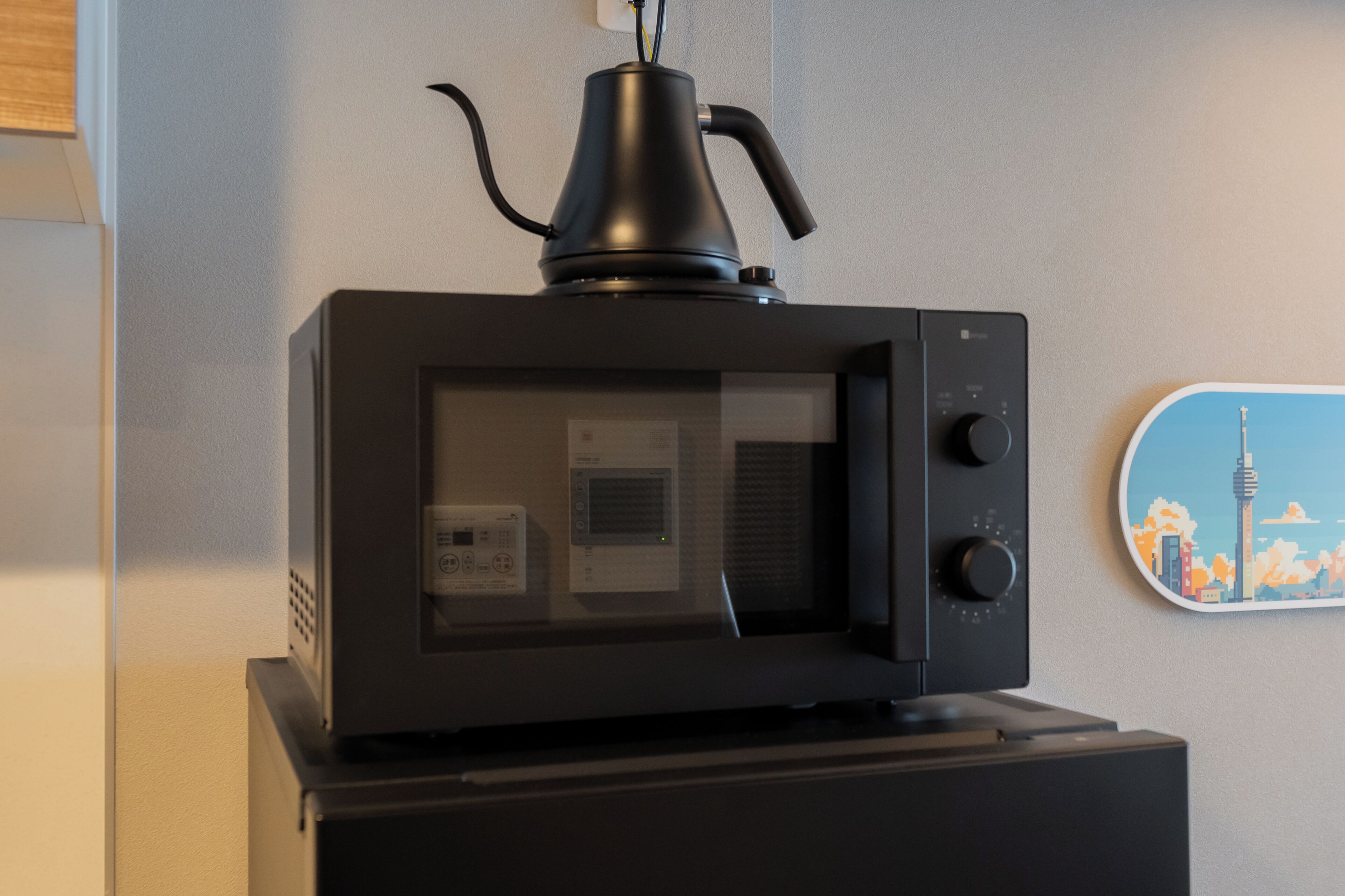 A microwave with a kettle on top, a small control panel, and a wall-mounted picture.