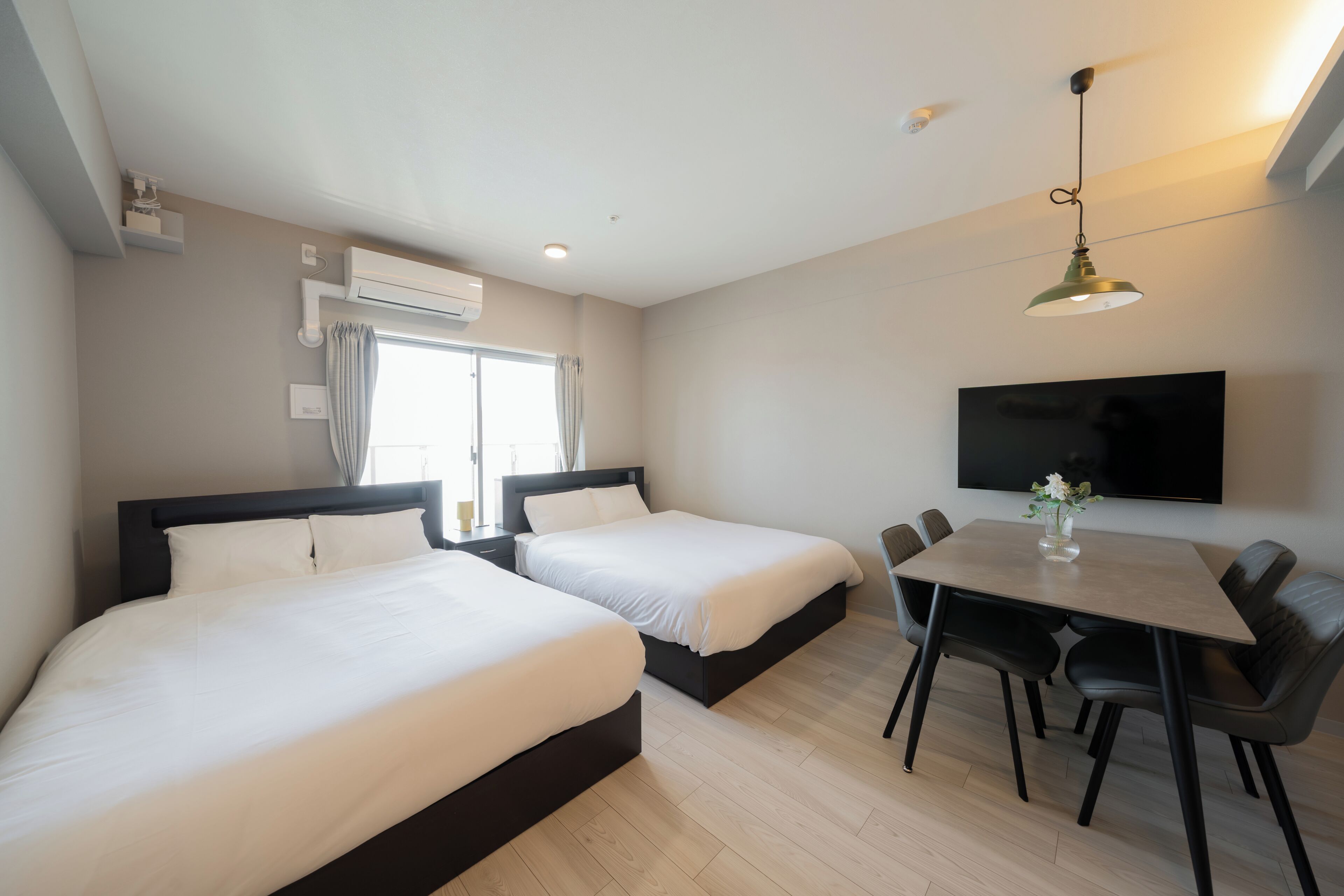 Une chambre d’hôtel avec deux lits, une table à manger avec des chaises, un téléviseur fixé au mur et un appareil de climatisation.