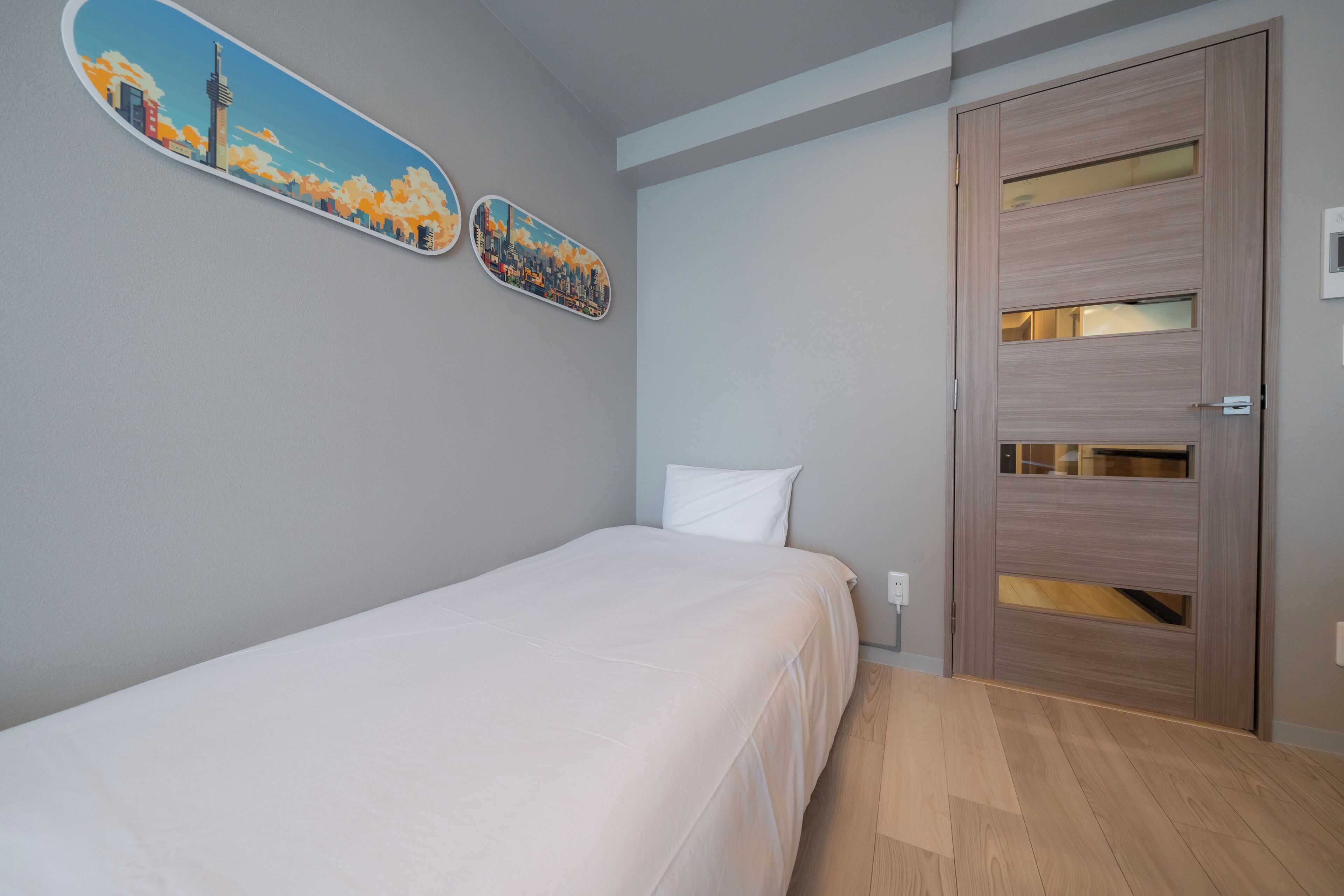 Un lit simple avec un matelas et un oreiller blancs, une porte en bois avec des panneaux de verre, et deux tableaux représentant des paysages urbains accrochés au mur.