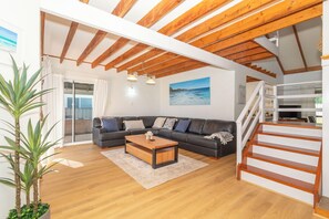 Een ruime woonkamer met een houten plafond, een zithoek, een salontafel, een trap en een groot raam.