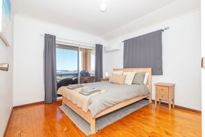 A bedroom with a wooden bed, a nightstand, a window with curtains, and a view of the outside. - JBay Hideaway: 4  BR, 2  BA House in Jurien Bay, Sleeps 8 (Jurien Bay)