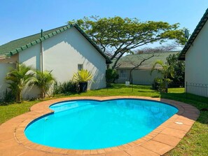 A swimming pool with a curved edge, surrounded by a brick deck. There are two buildings with green roofs and a tree in the background. - Coastal Chic in the Heart of Ballito 3-bed Oasis (Ballito)