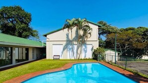 A swimming pool with a brick border, a building with a green roof, and palm trees. - Coastal Chic in the Heart of Ballito 3-bed Oasis (Ballito)