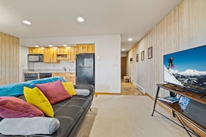 A modern living room with a black leather sofa, colorful cushions, a flat-screen TV on a wooden stand, and a kitchen in the background. - New! Affordable Mountain Escape | Pool - Hot Tub | Near Mt. Bachelor (Bend)