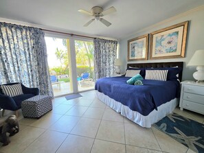 A bedroom with a bed, a nightstand, a chair, a sofa, a ceiling fan, and a view of the outdoors. - Sapphire 117 - 2 Bedroom Condo at Sapphire Beach on Dover Beach Barbados (Bridgetown)