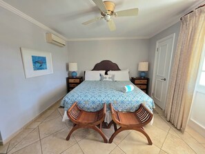 Une chambre à coucher comprenant un lit, deux tables de chevet, un ventilateur de plafond, un tableau encadré et une fenêtre avec des rideaux.