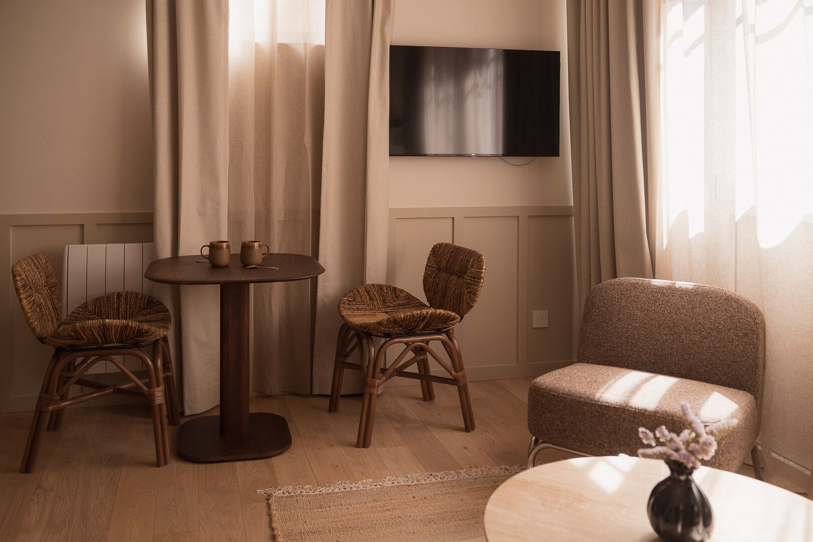 A room with a round table, two wicker chairs, a sofa, a TV mounted on the wall, and a vase with flowers on a side table.