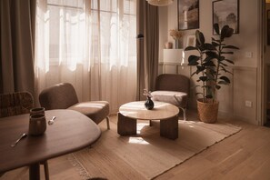 Una sala de estar con una mesa redonda, dos sillones, un sofá, una planta en maceta y un florero con flores.