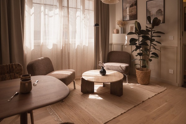 Una sala de estar con una mesa redonda, dos sillones, un sofá, una planta en maceta y un florero con flores.
