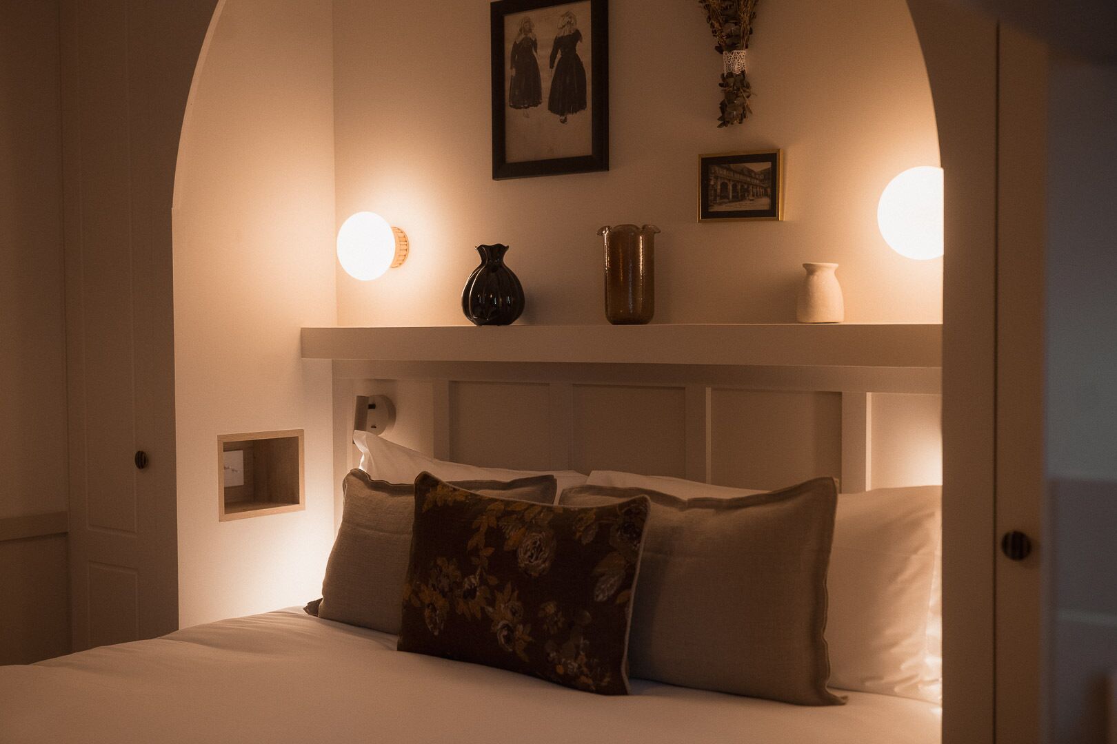 A bedroom with a bed, pillows, a nightstand, a wall-mounted shelf with decorative items, and wall sconces.