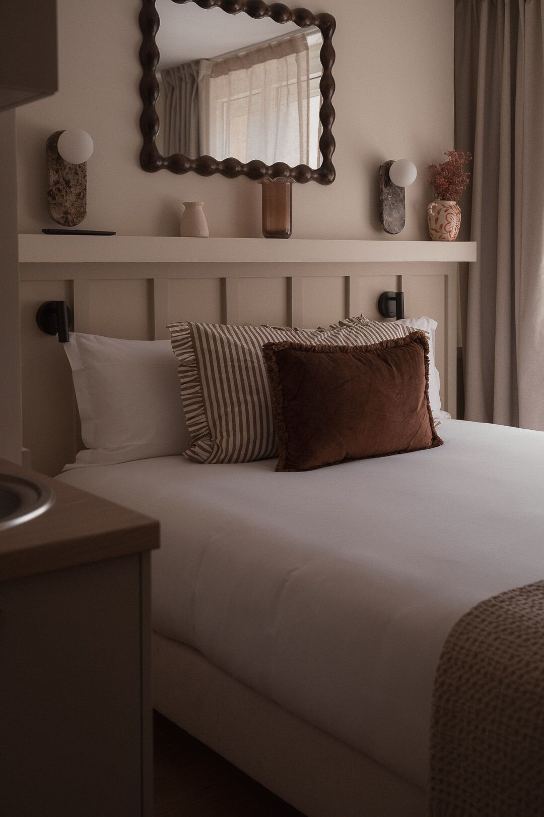 A neatly made bed with pillows, a bedside table, a mirror, and a shelf with decorative items.