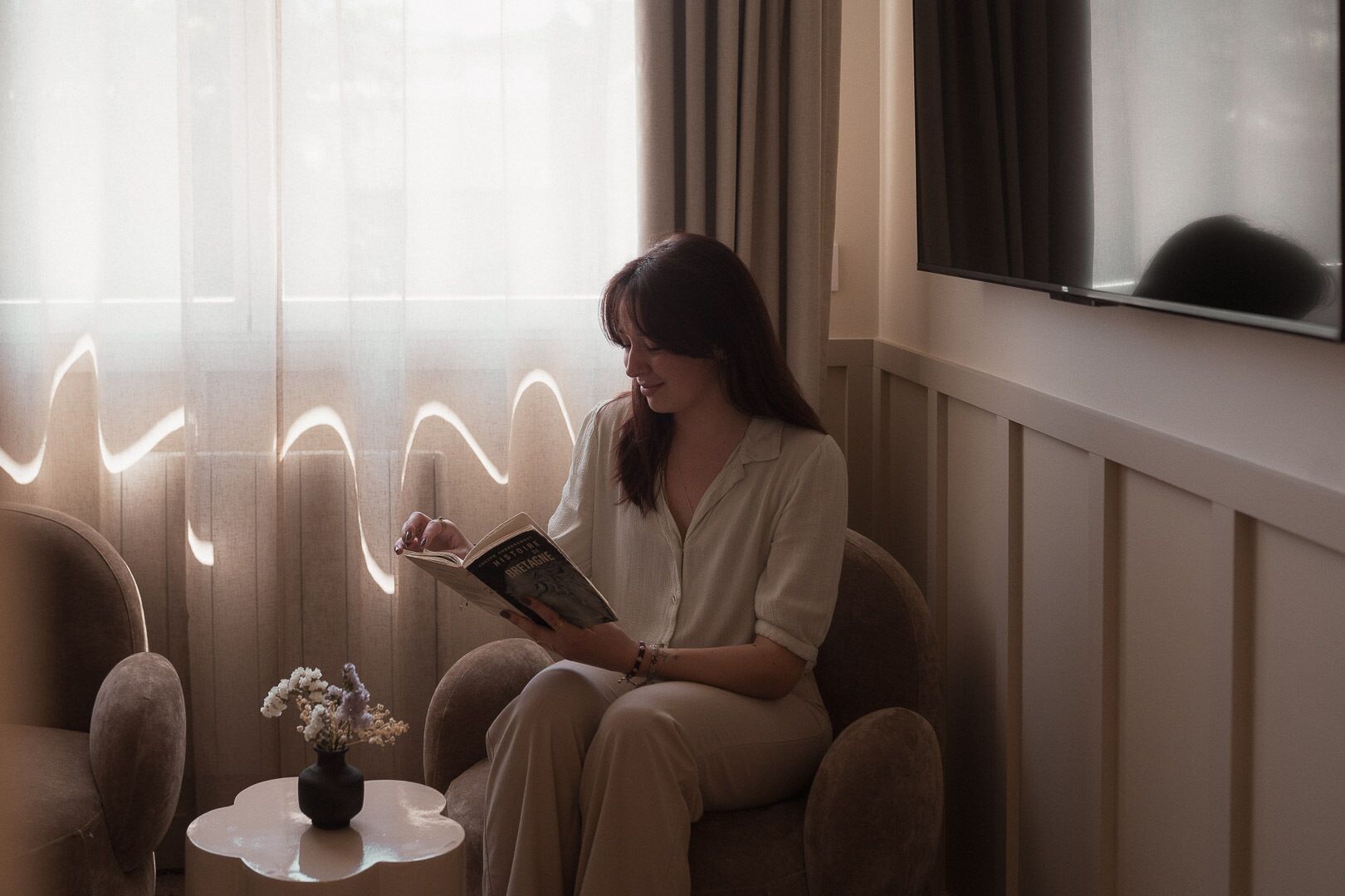Une personne est assise sur une chaise, lisant un livre dans une pièce qui contient une fenêtre, une petite table avec un vase et un miroir accroché au mur.