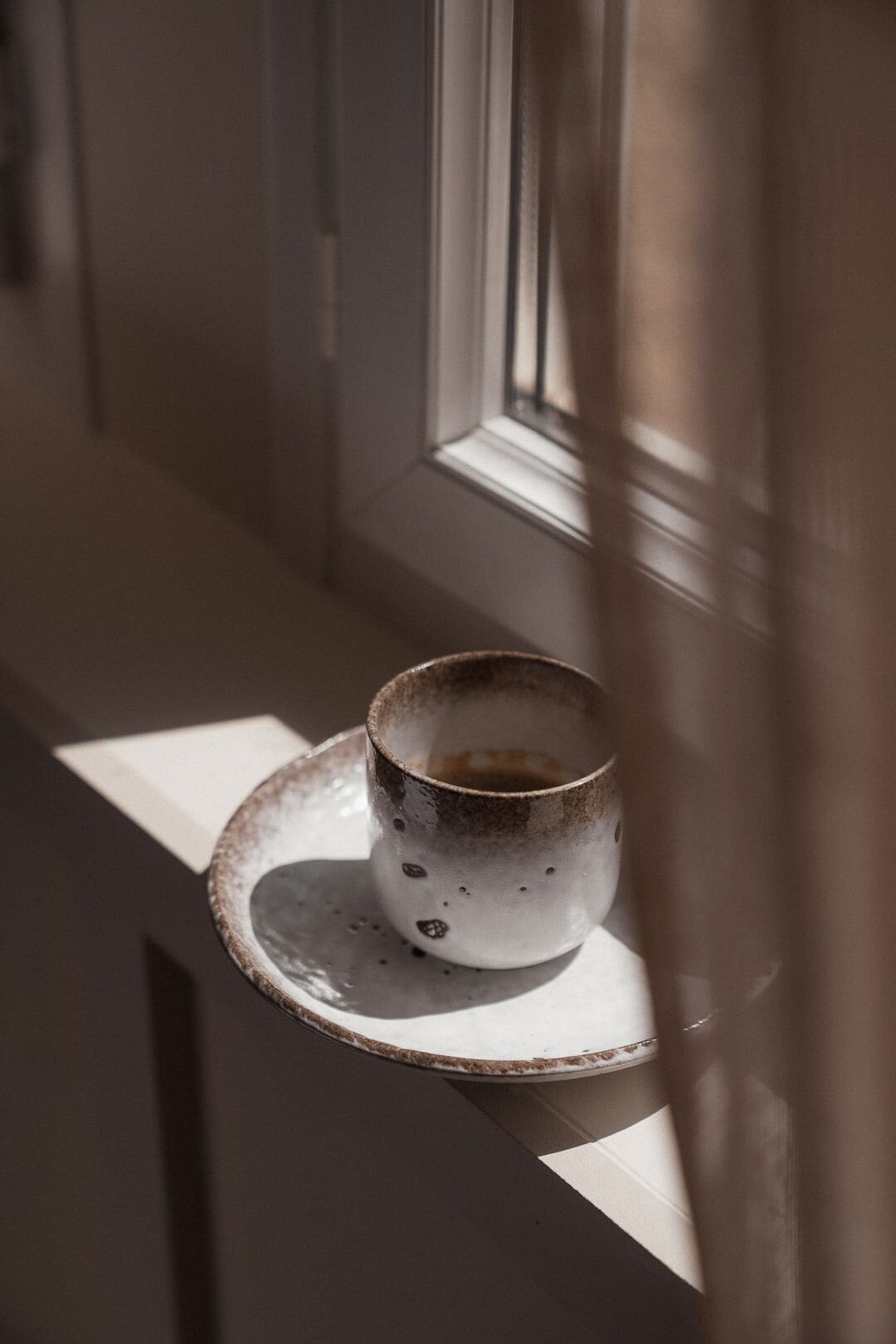 A cup on a saucer by a window with a curtain partially drawn.
