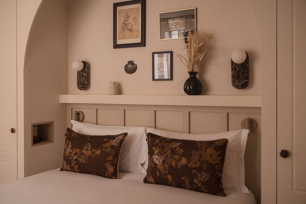 A bedroom with a bed, two pillows, a nightstand, a vase with dried plants, and framed pictures on the wall.