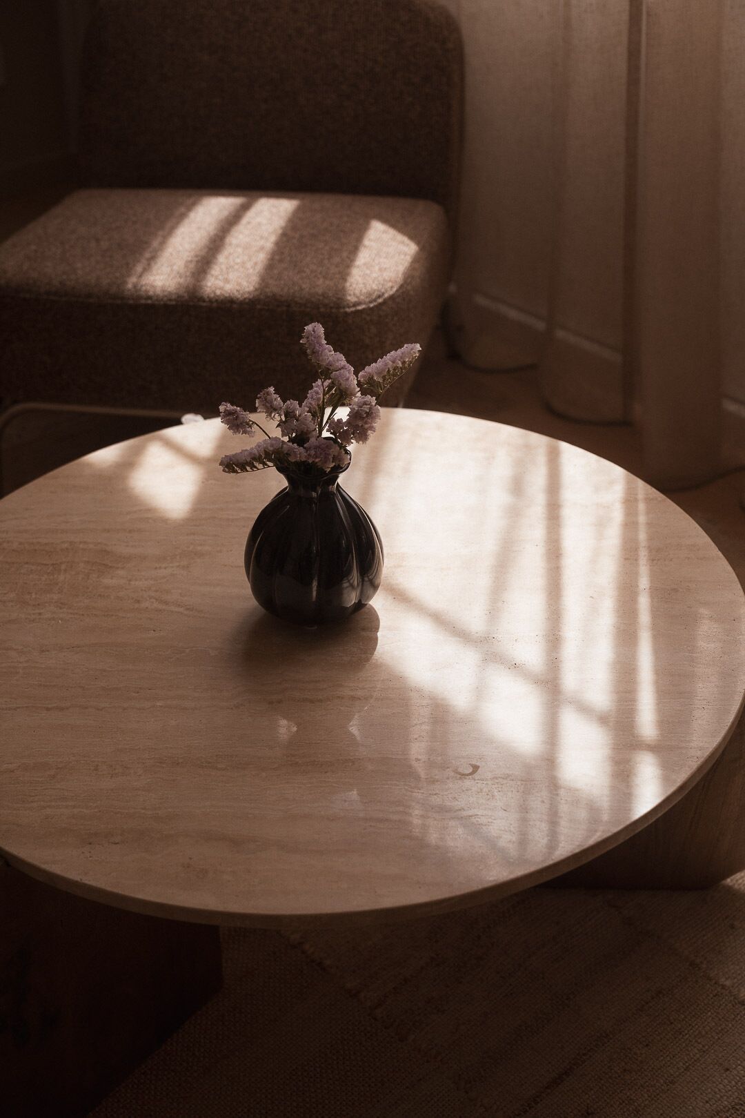 Une table ronde en bois, sur laquelle est posé un vase foncé contenant des fleurs, se trouve dans une pièce où l’on aperçoit un canapé marron et une fenêtre qui projette des ombres.