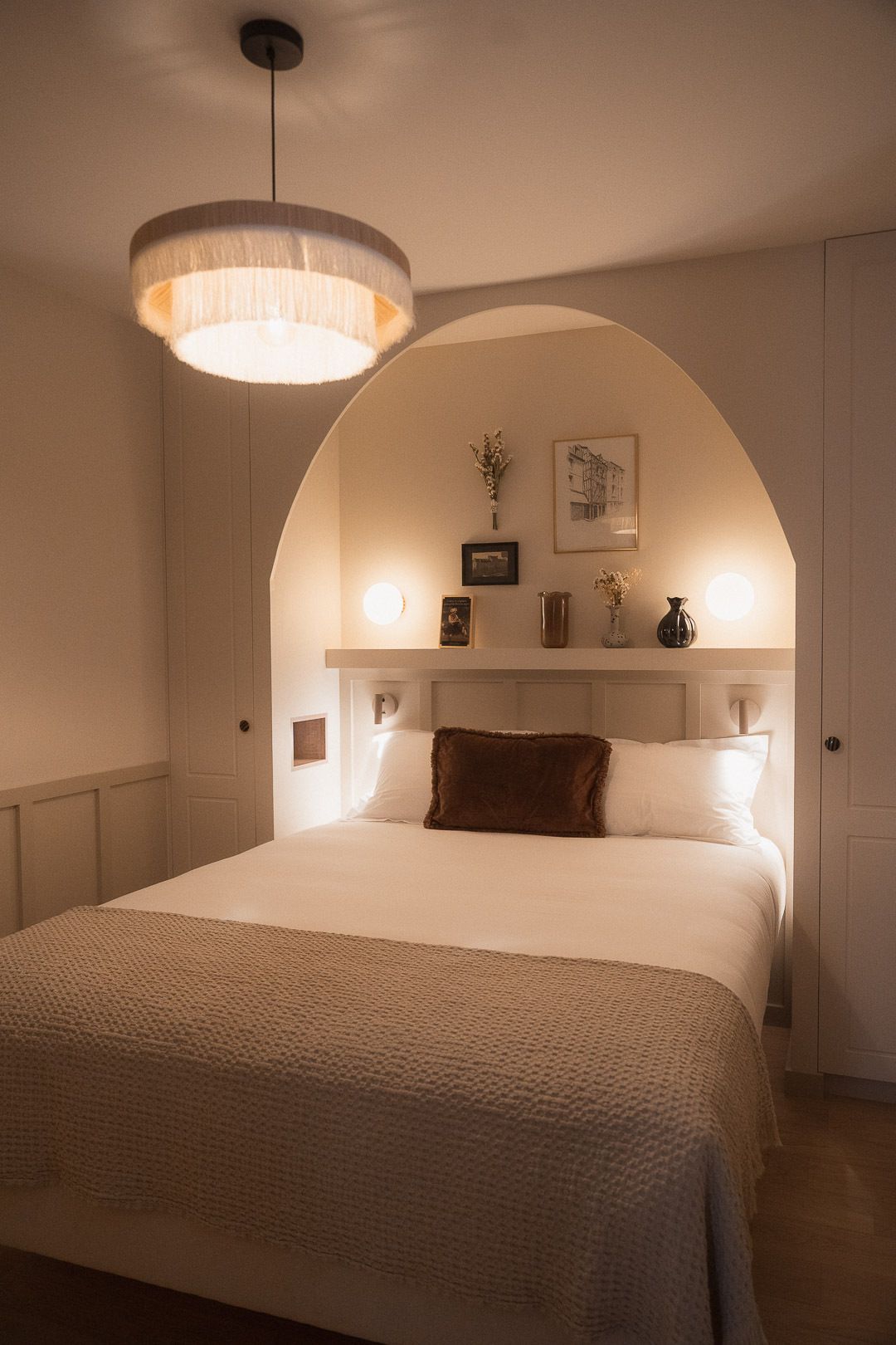 A bedroom with a large bed, an arched ceiling, a hanging light fixture, and a bedside table with a vase and picture frames.