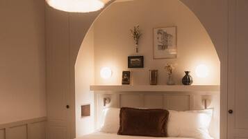 A bedroom with a large bed, an arched ceiling, a hanging light fixture, and a bedside table with a vase and picture frames.
