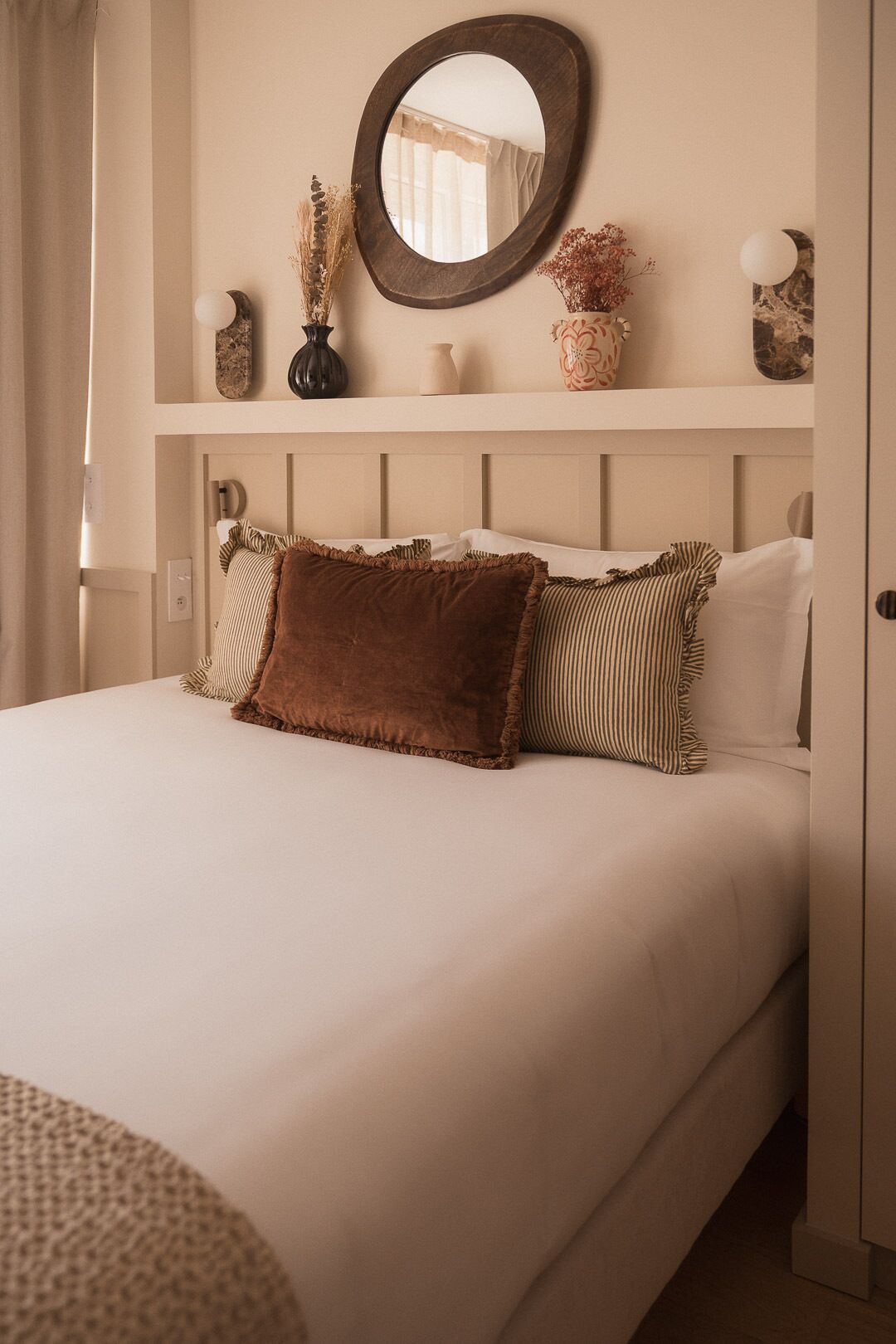 A bed with a headboard, pillows, a mirror, and decorative items on a shelf.
