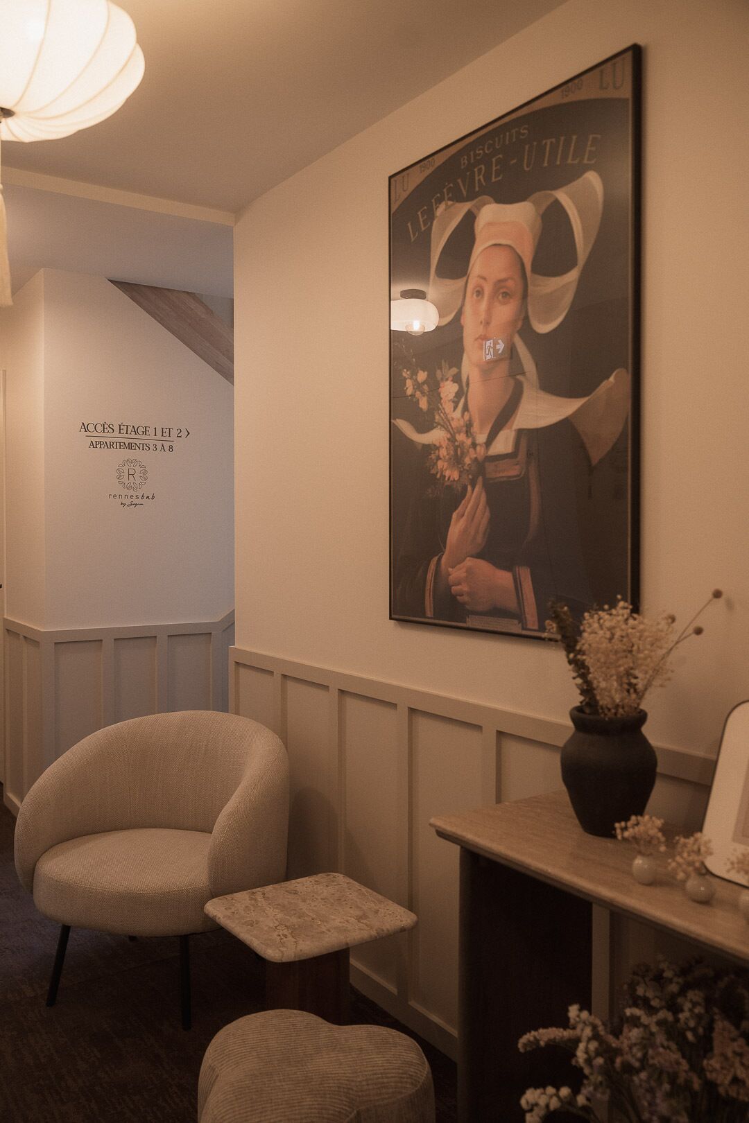 A hallway with a poster, a chair, a small table, and a vase with flowers.