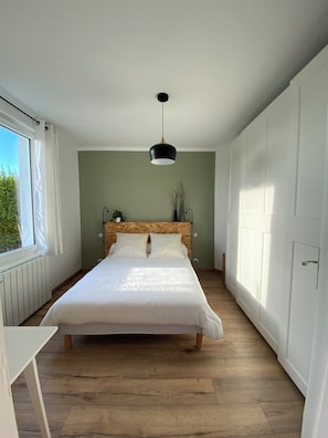 A bedroom with a wooden headboard, a bed with white bedding, a hanging pendant light, a radiator, and a window with white curtains. - 1930s house, ideal for families, close to beaches (Plounéour-Brignogan-plages)