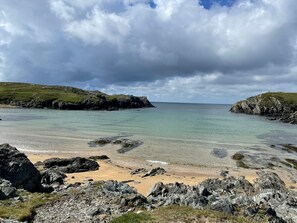 A coastal landscape with rocky terrain, a sandy beach, and a calm sea under a cloudy sky. - Goferydd (Holyhead)