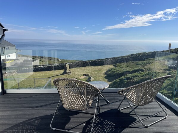A balcony with two wicker chairs and a small table, overlooking a coastal landscape with a stone wall and grassy area. - Goferydd (Holyhead)