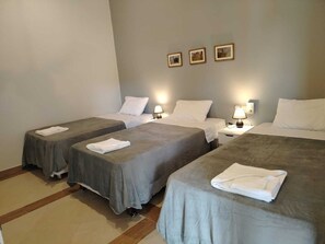 Quarto de hotel com três camas, cada uma com almofada branca e toalha branca dobrada. Há três abajures fixados na parede, e o chão do quarto é revestido de azulejos.