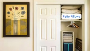 A room with a white door, a framed artwork on the wall, and a section labeled 'Patio Pillows' with neatly folded pillows and a hanger. - FIFA26 Chic Suite - Near NRG  (Houston)