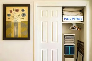 Ein Zimmer mit einer weißen Tür, einem gerahmten Kunstwerk an der Wand und einem Bereich mit der Aufschrift 'Patio Pillows', wo ordentlich gefaltete Kissen und ein Kleiderbügel ausgestellt sind.