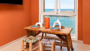 A room with a wooden table, two stools, a TV mounted on the wall, and a window with a view of the sea. - Waterfront Apartment - Ocean View (Essaouira)