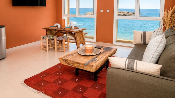 A living room with a TV, a table, chairs, a sofa, and large windows with a view of the sea. - Waterfront Apartment - Ocean View (Essaouira)