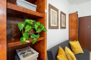 Ein hölzernes Bücherregal mit Büchern und einer Pflanze, ein graues Sofa mit gelben Kissen und zwei gerahmte Bilder an der Wand.