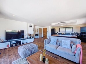 A modern living room with a flat-screen TV, a grey sofa, a wooden coffee table, and a kitchen area in the background. - Beautiful villa with private pool, 20 minutes from the beach and 10 minutes from the airport. (Sotta)