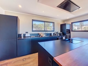 A modern kitchen with dark cabinetry, a central island, and two windows providing views. - Beautiful villa with private pool, 20 minutes from the beach and 10 minutes from the airport. (Sotta)
