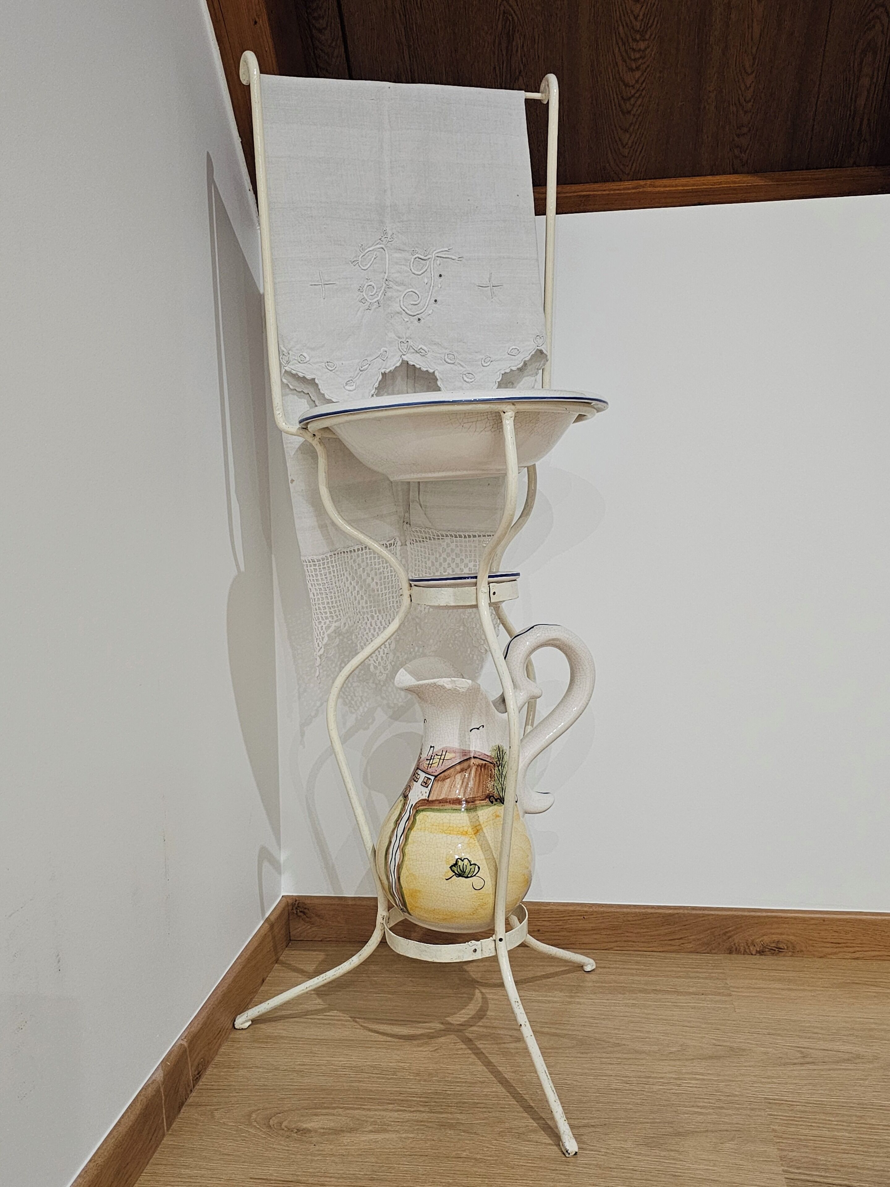 Una mesa de metal blanco con un florero y un mantel blanco encima, ubicada frente a una pared y piso de madera.