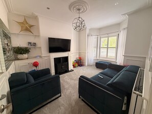 Ein Wohnzimmer mit dunkelblauen Sofas, einem Flachbildfernseher, einem Kamin und einem großen Fenster mit weißen Vorhängen.