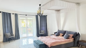Ein Schlafzimmer mit einem großen Bett, zwei Stühlen, einem Nachttisch, einer Kronleuchter und Blick durch ein Fenster mit Vorhängen.