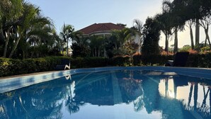 Ein klares Schwimmbecken mit gefliestem Rand, umgeben von Palmen und einem Gebäude mit rotem Dach im Hintergrund.