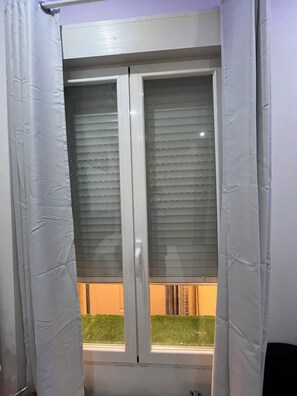 Ein Fenster mit horizontalen Rollläden, einem weißen Vorhang und Blick auf einen grünen Heckenstreifen.