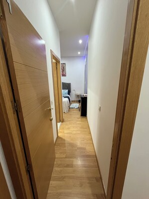 Un pasillo estrecho con suelo de madera, una cama con manta azul, una mesita pequeña y un cuadro colgado en la pared.